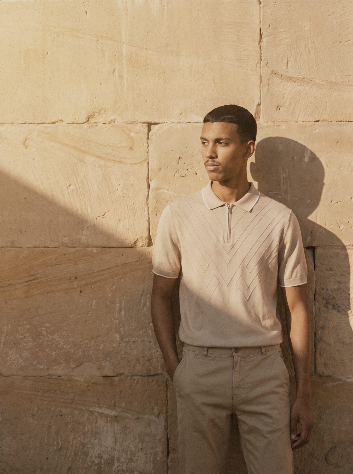 A man in a beige knit zip polo against a sandstone wall
