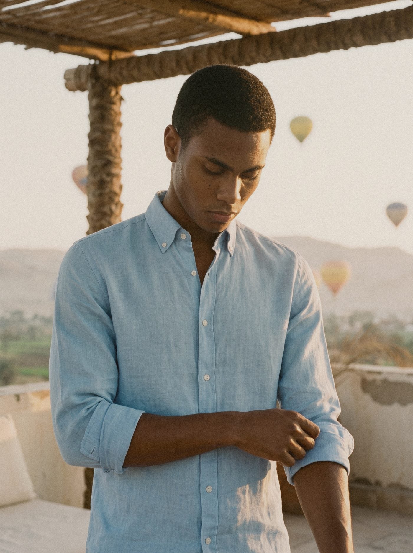 A man rolling the sleeve of a light blue linen shirt on a rooftop terrace at dawn