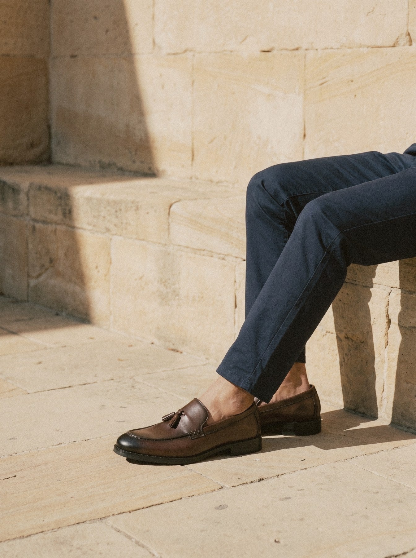 Brown tassel loafers and navy chino hems, seated on a sandstone ledge