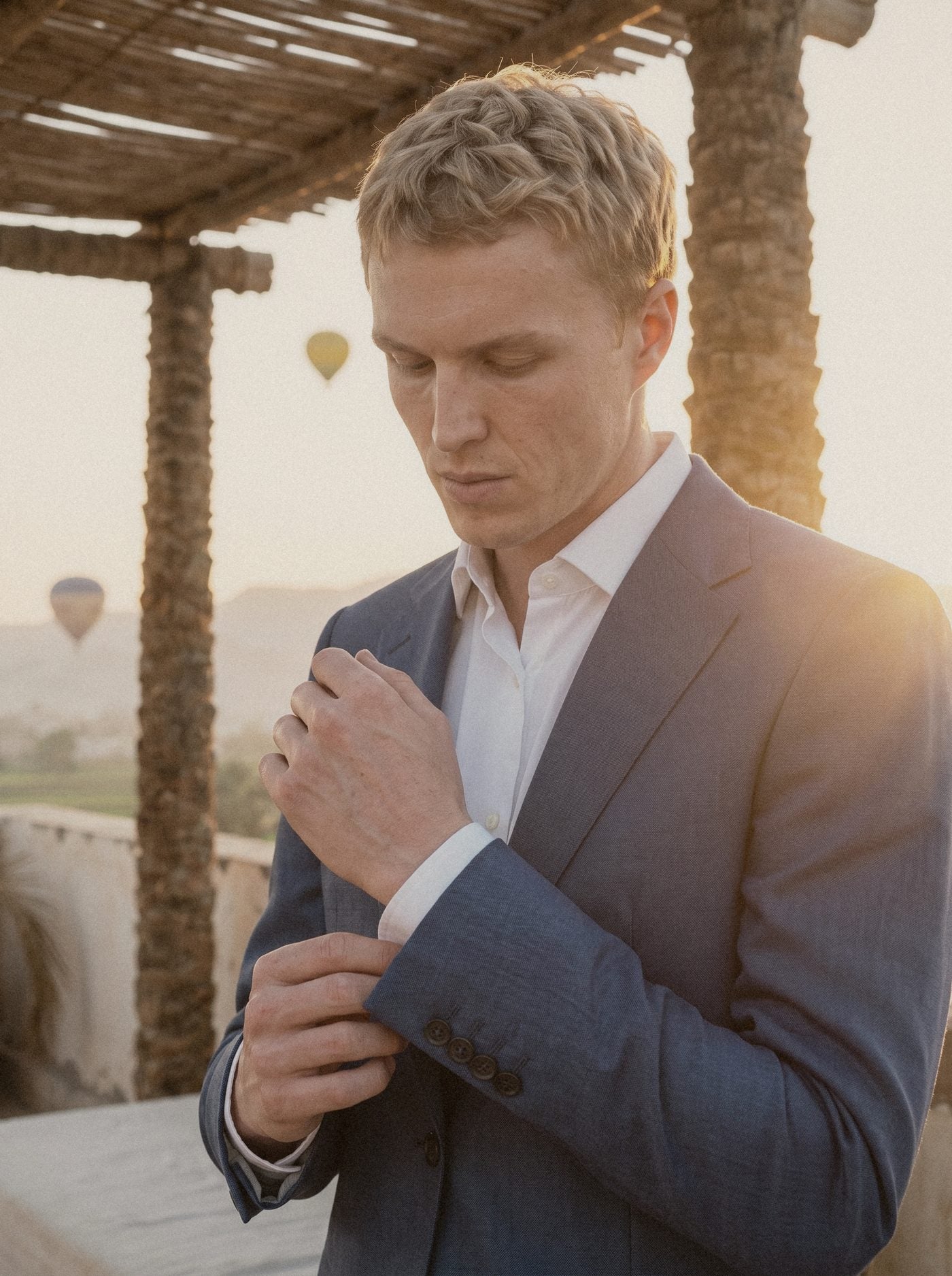 A man in a mid blue suit settling his cuff on a rooftop terrace at dawn