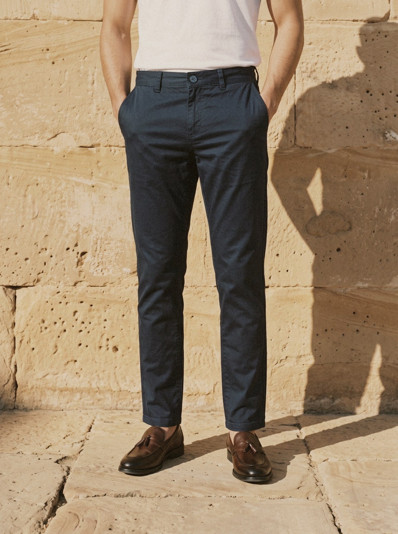 Navy cotton chino with a white tee tucked in, hands in the pockets, against a sandstone wall